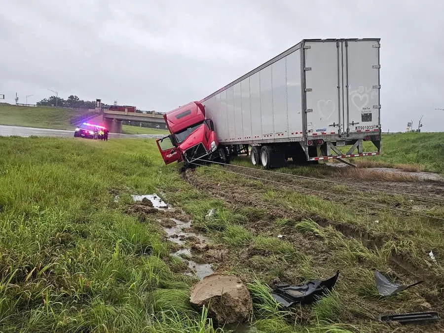 A red semi tractor and loaded dry van off the road in a wet ditch below an overpass, recovery lines rigged and a patrol car on the shoulder.